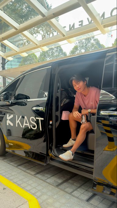 a young man wearing a pink shirt and white sneakers sitting in the open sliding door of a black van with 'K KAST' branding