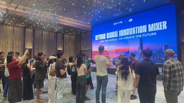 The image shows a large group of people gathered around a big screen displaying an advertisement or presentation related to global innovation. The attendees are standing close together, indicating that they have just attended an event like the Global Innovation Mixer held at the Shanghai World Expo in 2013. The crowd seems engaged and interested in what is being presented on the screen, which could be about new technologies, ideas, or initiatives aimed at driving innovation forward.