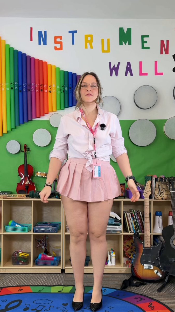 A woman in a pink skirt and white blouse stands confidently, holding a guitar on her shoulder, with a rainbow-colored wall and musical instruments behind her.