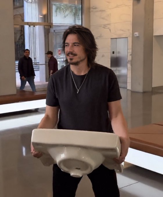 A man with long dark hair and a beard, wearing a dark t-shirt, stands in a modern building lobby while holding a white ceramic toilet bowl in front of him.