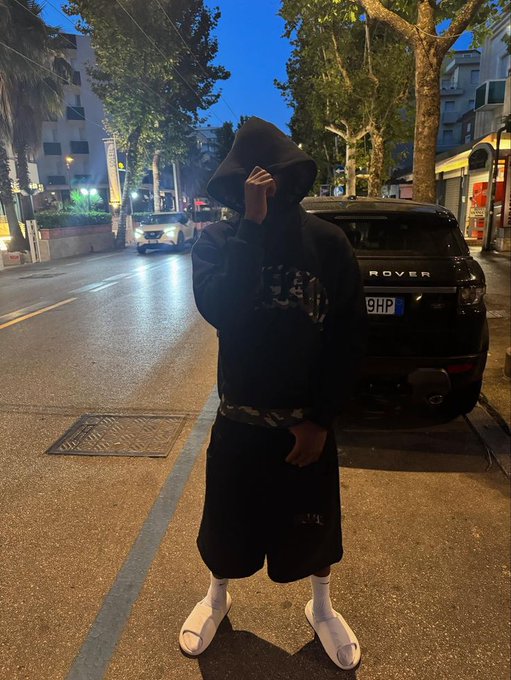 A person in a dark hoodie and shorts stands on a dimly lit street next to a Range Rover with a "9HP" license plate, embodying the late-night grind mentality referenced in the post. The hooded, anonymous look reinforces the "keep stacking" message, with the parked Rover serving as visual motivation for the continued hustle toward $River points.