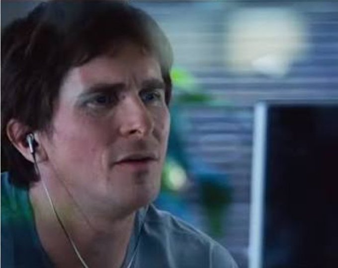 A close-up shot of a man with brown hair and earbuds, looking intensely off-camera in what appears to be an office setting.