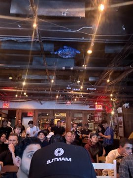 The image shows a crowded restaurant with people sitting at tables and enjoying their meals. A man wearing a black baseball cap stands out among the crowd, possibly taking a photo or capturing a moment from the event. The atmosphere appears lively and social as patrons gather together in this public space for dining and entertainment.