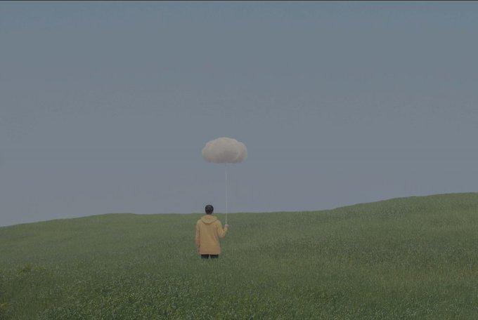 A person in a yellow jacket stands in a grassy field holding a string attached to a small white cloud balloon against a grey sky.