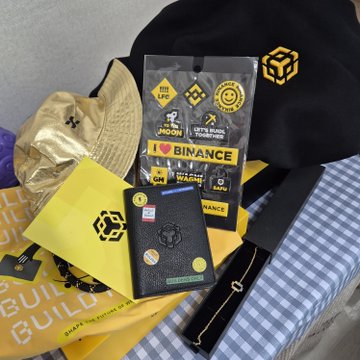 A collection of Binance-branded merchandise and stickers including a black hat, gold cap, sticker pack with "BINANCE BUIDL" and "LET'S BUILD" text, wallet, and other promotional items arranged on a blue checkered surface.