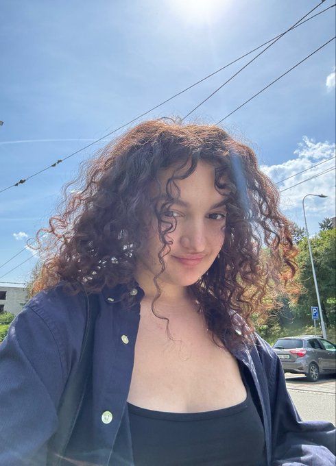 Outdoor selfie of a young woman with curly brown hair wearing a black top and an open dark blue button-up shirt, taken on a sunny day with power lines and a car visible in the background.