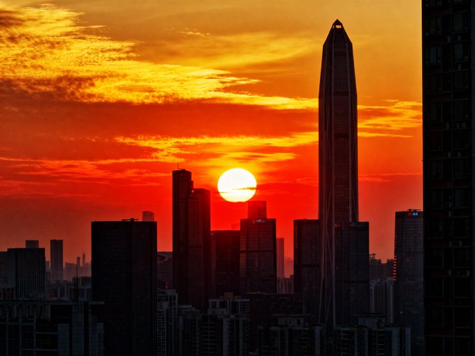 A dramatic sunset cityscape silhouette showing the sun setting behind tall skyscrapers, with a vibrant orange and red sky, likely depicting a major Asian metropolitan skyline.