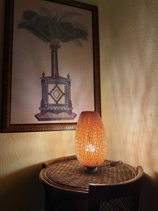 An illuminated orange decorative lamp with a diamond pattern sits on a wicker side table, with a framed black-and-white photo of a palm tree and pedestal hanging on the wall behind it.