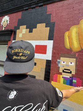 A person wearing a 'BORED APE YACHT CLUB' cap and matching black t-shirt standing in front of a colorful mural featuring pixel-art characters.