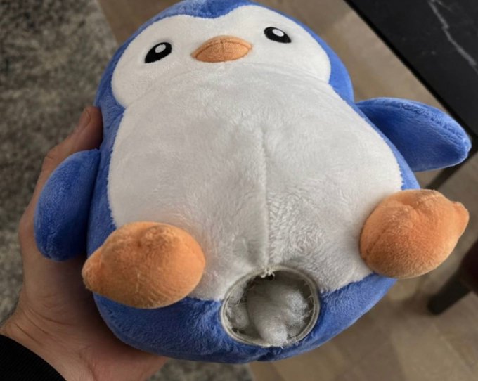 A hand holds a blue and white plush penguin toy that appears to have a tear or opening on its belly, revealing the white stuffing inside.