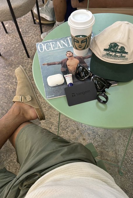 A casual outdoor scene showing a person's legs and feet resting near a green table holding a Starbucks coffee, an 'OCEAN' magazine, a cap, sunglasses, AirPods, lip balm, and a black 'tangem' card.