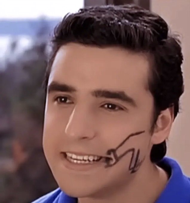 A close-up portrait of a smiling young man with dark hair, wearing a blue shirt, with a stylized letter 'N' drawn or projected on his cheek.