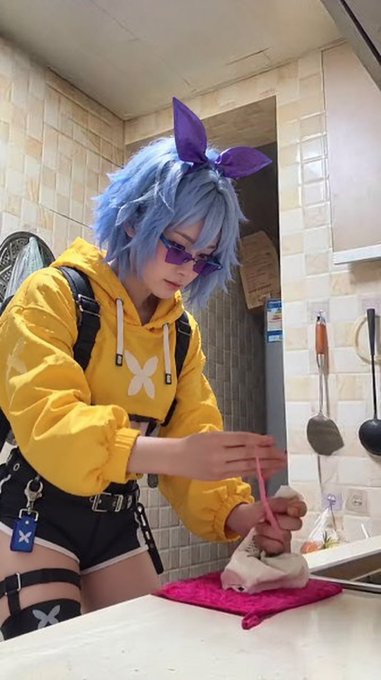 A cosplayer with blue hair and purple bunny ears, wearing a yellow hoodie and black shorts, is focused on doing dishes at a kitchen sink, fitting the post's humorous nod to prepping for upcoming "$FLAPA" announcements.