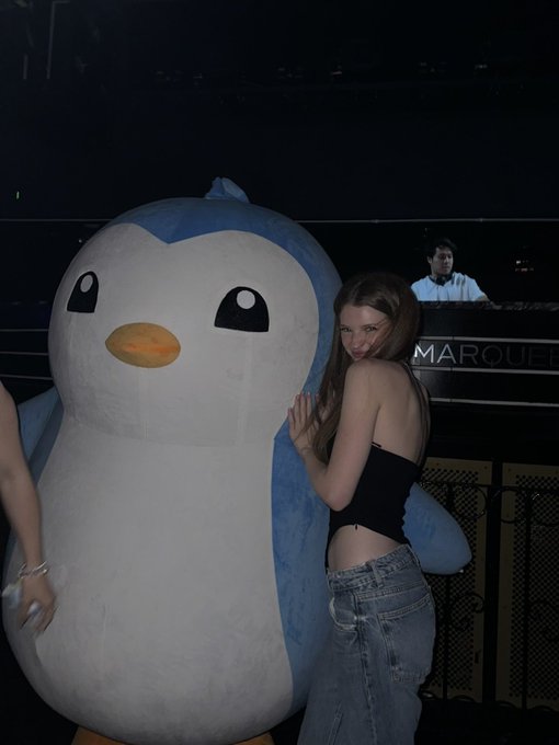 A young woman with long brown hair poses in a black tank top and jeans, hugging a large blue and white penguin mascot at what appears to be a nightclub or event venue.