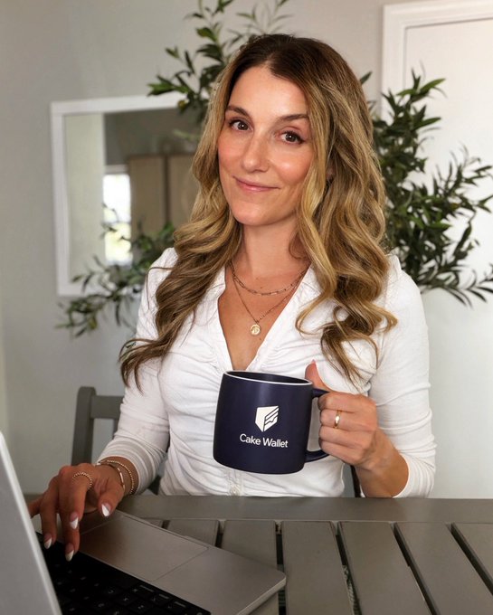 A woman with long wavy blonde hair sits at an outdoor table working on a laptop while holding a navy blue mug that reads 'Cake Wallet'.