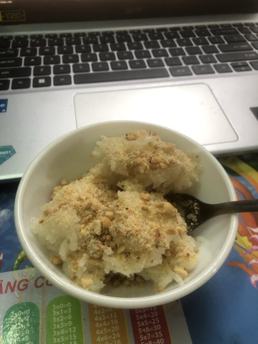 A bowl of sticky rice topped with crushed peanuts sits on a colorful children's multiplication table mat in front of an open laptop keyboard.
