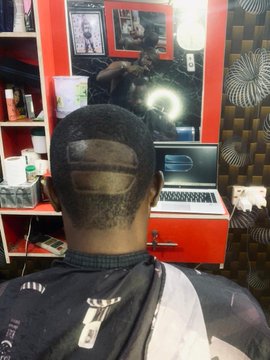 Photo showing the back of a person's head with a distinctive geometric hair design shaved into their hair, sitting in front of a red-framed mirror in what appears to be a barbershop or salon.