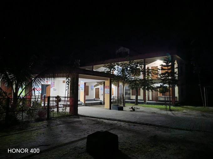 A nighttime photograph taken with an Honor 400 (watermark visible) showing a two-story building with an illuminated entrance and porch, surrounded by trees and palm trees against a dark sky.