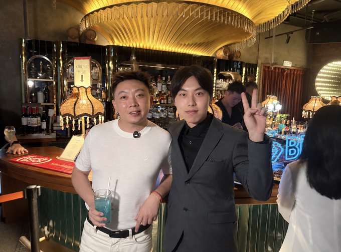Two men pose together in a stylish bar with ornate lighting; the man on the left holds a blue drink while the man on the right flashes a peace sign in a gray suit.