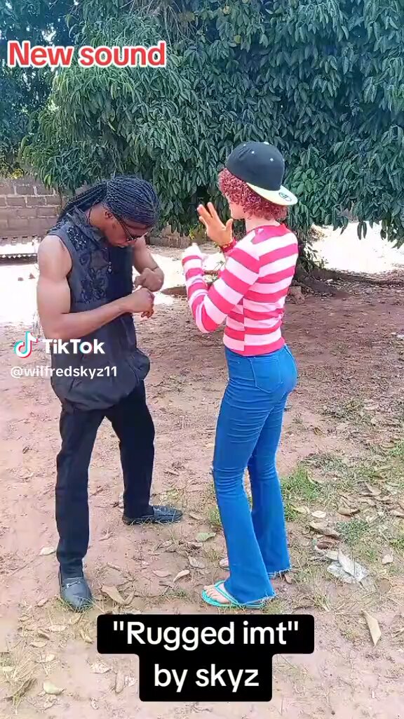 The image features a man and woman standing next to each other, with the woman wearing a pink shirt and blue jeans while the man has long hair and is dressed in black. The background includes trees and bushes, suggesting an outdoor setting. There are also text elements within the image that read "New Sound" and "Monad isn' t dead one bit".