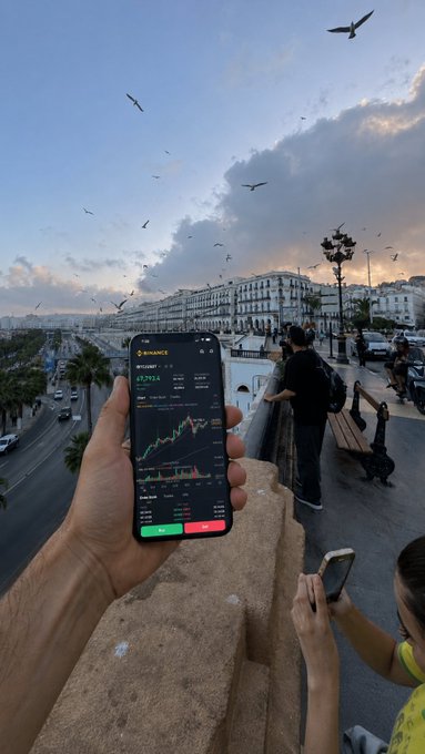 A hand holds up a Binance app displaying the BTC/USDT chart showing a price of 67,974.4, with a scenic coastal promenade, birds in the sky, and Mediterranean-style architecture visible in the background—hinting at the post's "Where do you think I am?" question by suggesting an exotic travel destination while crypto trading on the go.