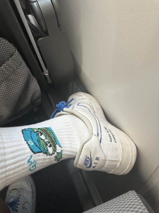 A casual photo taken inside a vehicle showing a foot resting on the dashboard, wearing white Nike Air sneakers and white socks featuring a cartoon alligator graphic.