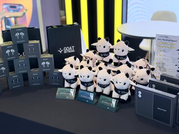 A promotional table at the GOAT Network booth displays stacks of Samsung Galaxy Buds boxes alongside plush goat mascots and Starbucks gift cards, with an information sheet reading "Meet The GOAT Agent" visible in the background. The setup aligns with the post's invitation to visit booth 37 at KBW, where attendees can engage with agent commerce and take home branded merchandise like the goat plushies shown.