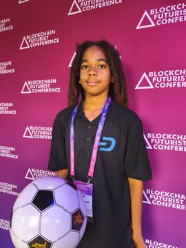 Young man with dreadlocks wearing a lanyard standing in front of a 'Blockchain Futurists Conference' backdrop while holding a soccer ball.