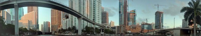 A panoramic cityscape shows an elevated train track curving through a modern downtown area with tall skyscrapers under a clear sky.