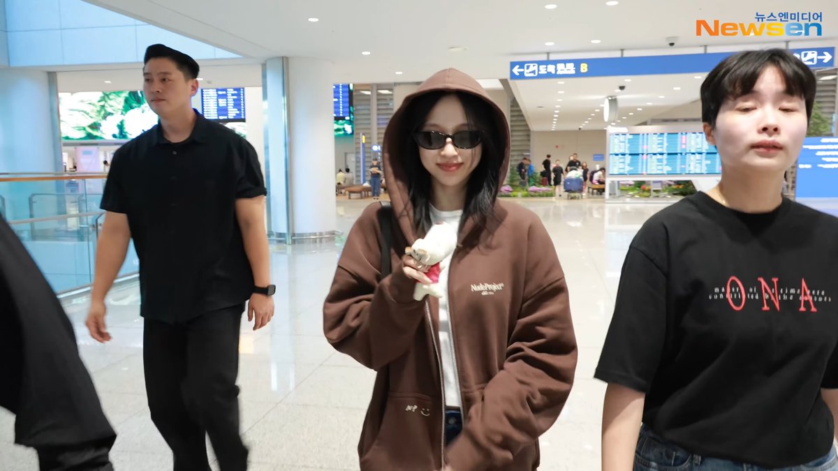 A young woman wearing a brown "NubProject" hoodie and sunglasses walks through what appears to be an airport terminal, flanked by two men in black shirts. The "Newsen" watermark and Korean signage indicating flight information in the background suggest this is her arrival back in Korea, consistent with the post about MINA returning from Indonesia after the SK-II event.