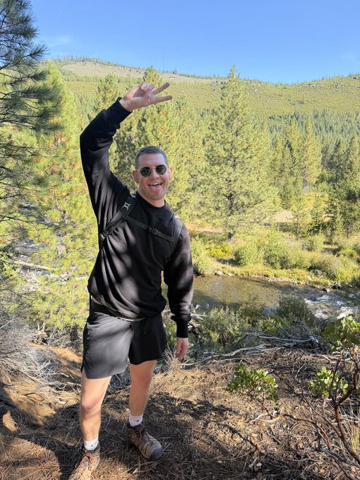 A man in a black sweatshirt, shorts, and sunglasses poses with a peace sign while hiking in a lush, mountainous area with a stream, playfully illustrating the "touching grass" theme.