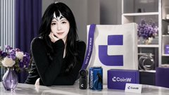 A low-resolution image of a smiling woman with dark hair sitting at a table next to a Coinbase promotional display showing 'Coinbase' text and a purple logo.
