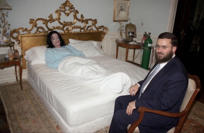 A woman rests in a luxurious bedroom with an ornate gold headboard while a man in a dark suit and yarmulke sits in a chair beside her, with an oxygen tank visible in the background—suggesting the caption "That's not..." refers to the unusual presence of an Orthodox rabbi appearing to be in a bedroom with a woman who isn't his wife.