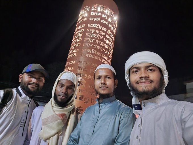 Four young men taking a selfie at night in front of a tall illuminated minaret covered in Arabic or Persian script, with the men wearing traditional Islamic caps and clothing.