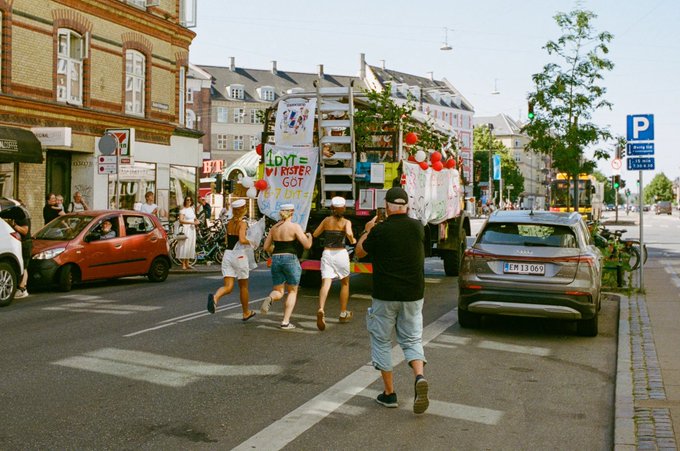 A parade truck with a banner reading 'LOVE NEVER GOT TOG' drives down a European street, followed by people in white sailor hats and shorts.