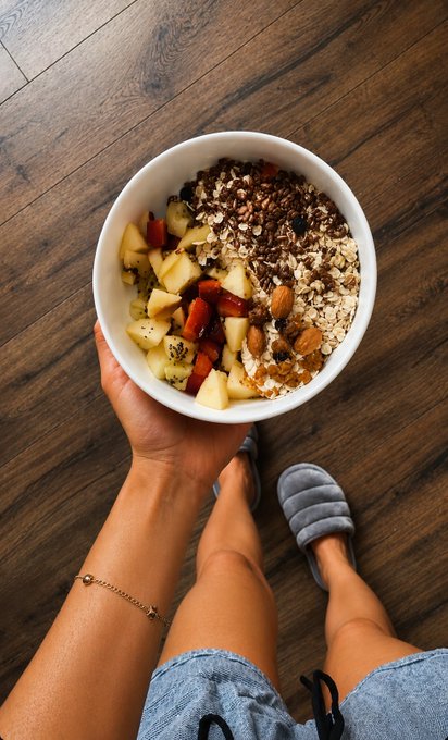 A first-person view shows a hand holding a white bowl filled with granola, almonds, kiwi, and fruit, with a leg wearing an anklet and gray slippers visible below.