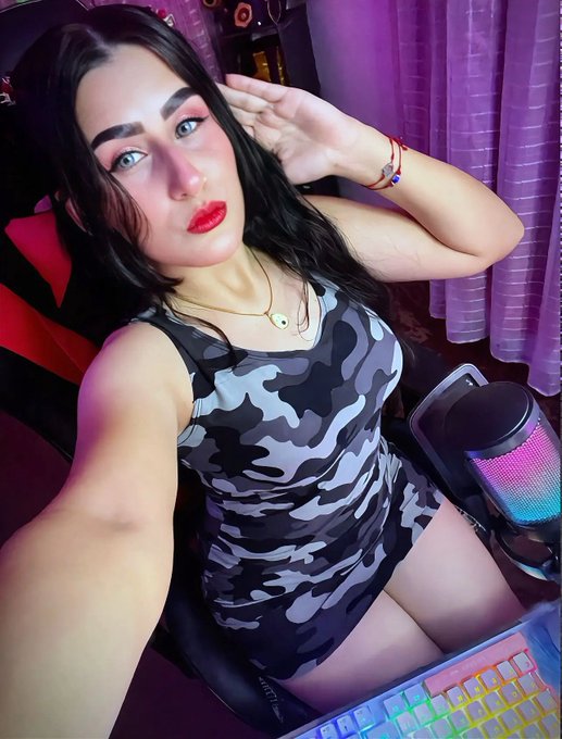 A woman with dark hair and bright red lipstick wears a black and gray camouflage dress, posing for a selfie in a gaming chair beside a colorful RGB-lit keyboard and microphone setup.