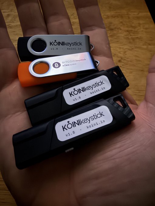 A hand holds four KoinKeystick hardware wallets labeled 'v1.0 koin1.io', with two featuring Bitcoin branding.