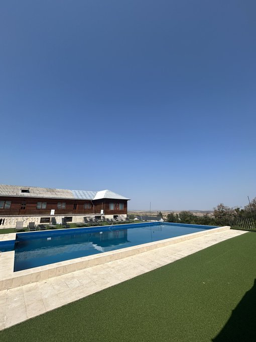 A bright sunny day showing a clear blue swimming pool surrounded by stone tiles and artificial turf, with a building and distant landscape in the background.