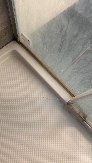 A bathroom shower floor corner showing water stains, soap residue, and a scuffed white shower base next to a glass door with blue-tiled walls.