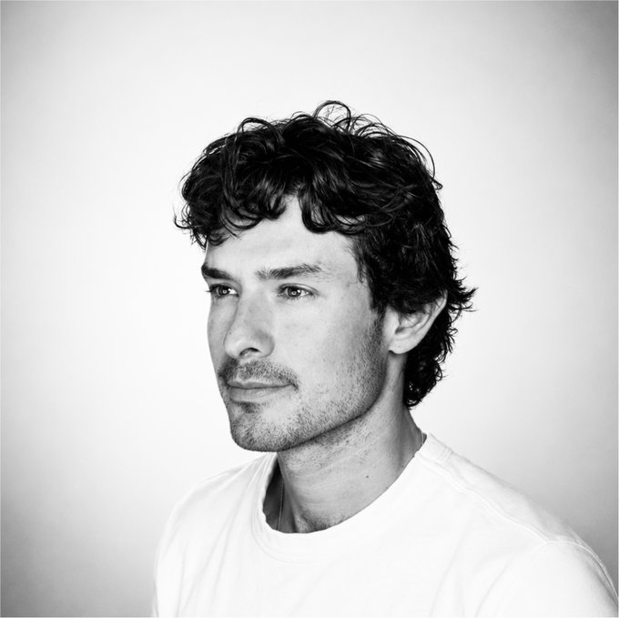 A black and white portrait photograph of a young man with dark curly hair, light stubble, and a white t-shirt, gazing thoughtfully to the side.