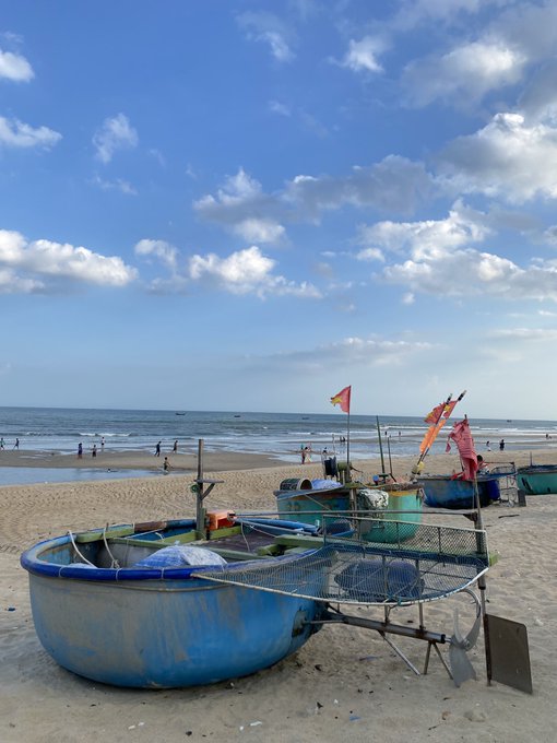 A serene beach scene with traditional round Vietnamese basket boats resting on the sand, colorful fishing flags fluttering in the breeze, and a bright blue sky filled with scattered clouds over the ocean.