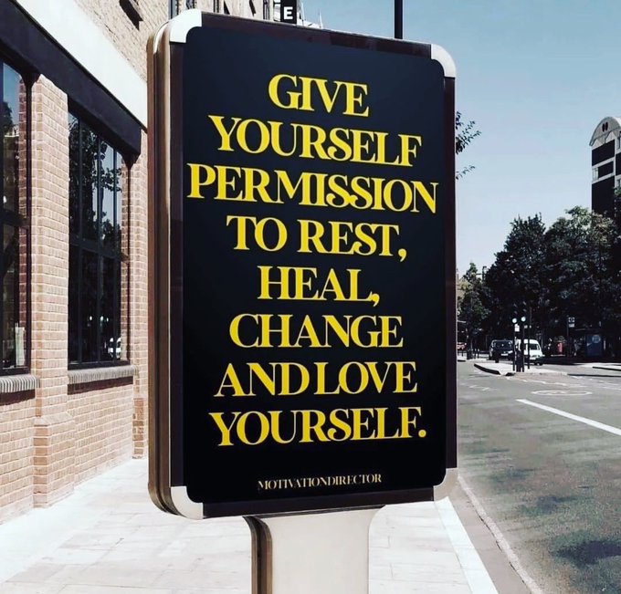 A black sign with yellow text on a city street reading: "GIVE YOURSELF PERMISSION TO REST, HEAL, CHANGE AND LOVE YOURSELF." with "MOTIVATIONDIRECTOR" below.