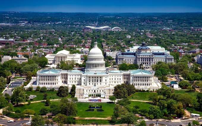 An aerial daytime view of the United States Capitol building in Washington D.C., showing the iconic white dome surrounded by green lawns and trees.
