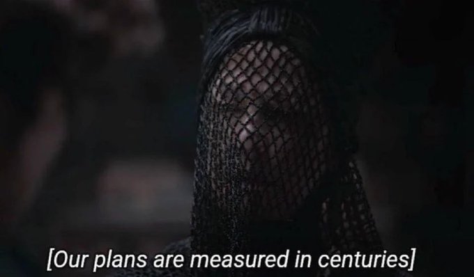A dark, mysterious figure is veiled in black netting with the subtitle '[Our plans are measured in centuries]' displayed at the bottom.