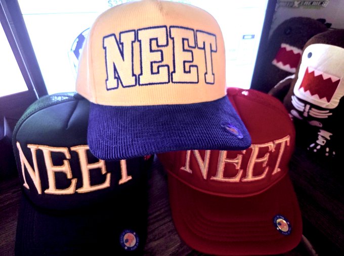 A collection of three baseball caps (cream, dark blue, and red) displayed with the word 'NEET' prominently embroidered in large block letters on each.