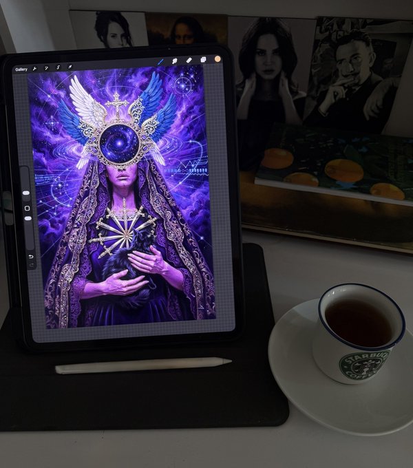 An iPad displays a mystical purple digital artwork of a winged figure with a celestial eye for a head, resting on a desk beside a cup of tea with 'Starbucks Coffee' visible on the saucer.