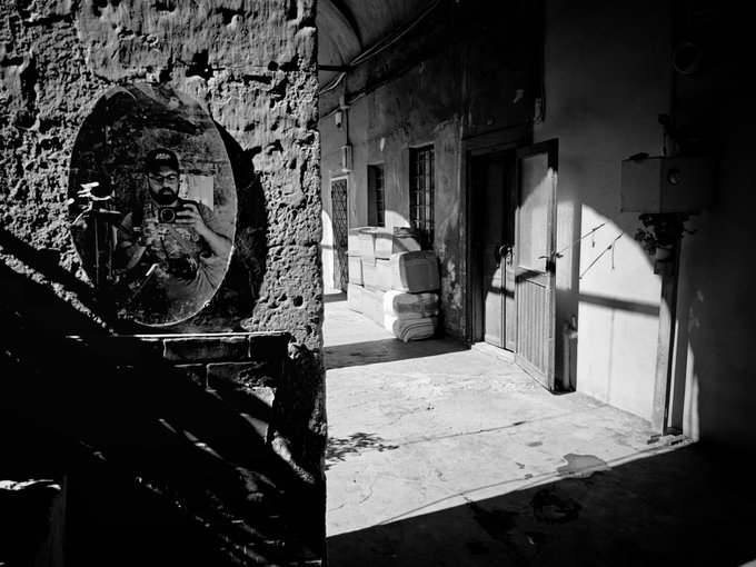 A black and white photograph showing a man taking a photo with his phone, reflected in a round mirror mounted on a stone wall, with a sunlit courtyard, stairs, and an open wooden door in the background.