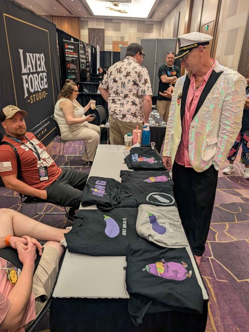 At a "Layer Forge Studio" booth, a man in a sparkling silver jacket and captain's hat browses a table displaying merchandise featuring "Just Blast" eggplant logos. The setup showcases branded apparel available at Dongarena.me, where fans can pick up their own gear.