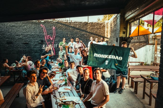The image shows a large group of people gathered around a long dining table, enjoying their time together. Some are sitting at the table while others stand nearby, creating an atmosphere of camaraderie and shared experiences. The scene takes place outdoors, possibly in a park or similar public space. A green banner is prominently displayed on the wall behind the group, which reads "Monkkedao". This suggests that the gathering could be related to a coworking event or a social gathering for web3 enthusiasts.